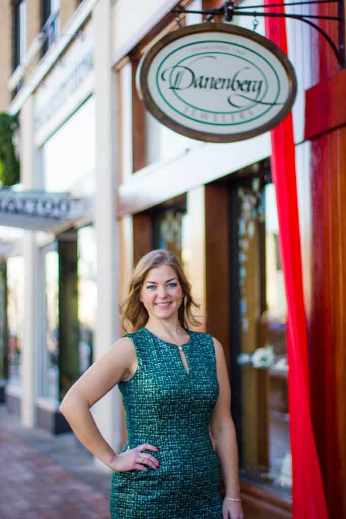 A woman in a green dress stands smiling with one hand on her hip in front of a storefront. Above her, an oval sign reads "Darenberg" in elegant script. The store has large windows and red accents. The street scene includes another store in the background.