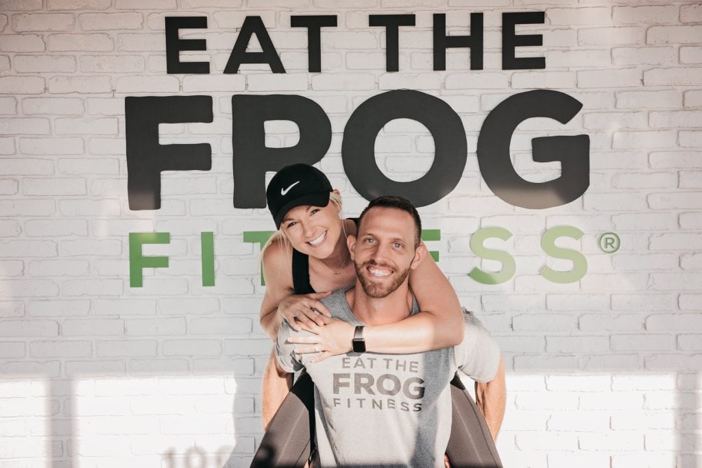 A smiling man and woman pose in front of a white brick wall with the words "EAT THE FROG FITNESS." The woman, wearing a black cap, hugs the man from behind, who is seated and also smiling. They both wear casual workout attire with the same logo.