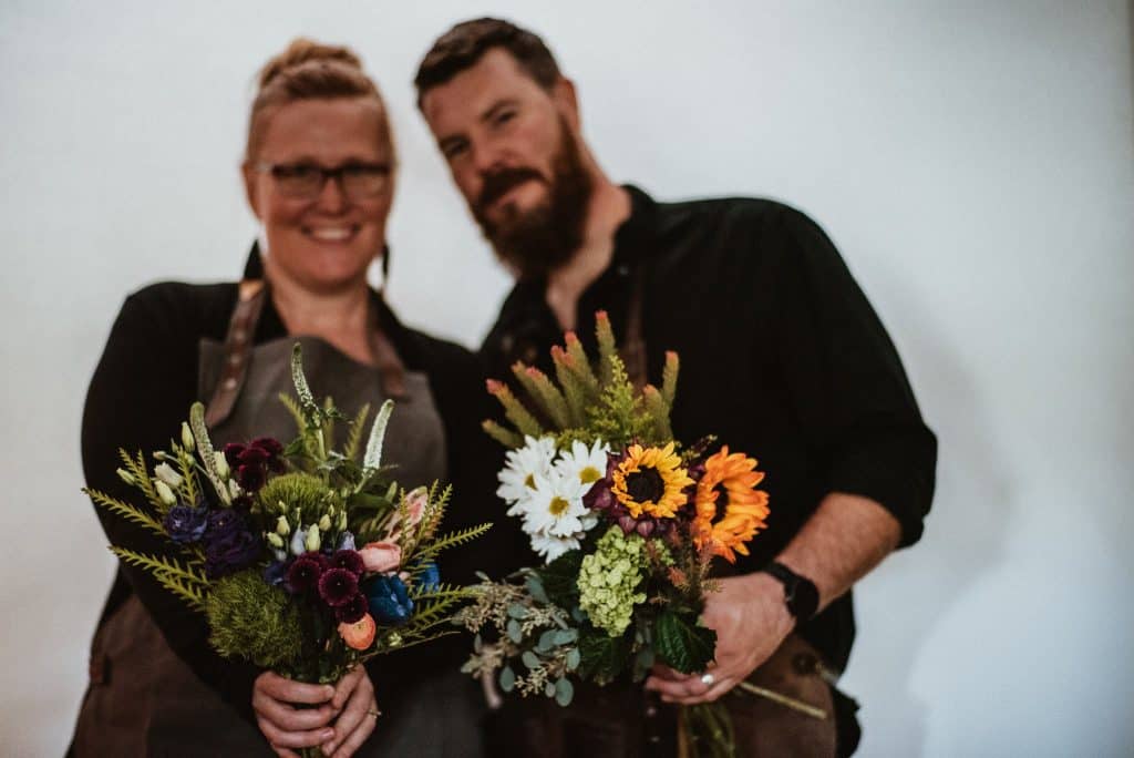 Two people are standing side by side, each holding a bouquet of flowers. The person on the left is wearing glasses and an apron, and the person on the right has a beard and is dressed in a dark shirt. They both appear to be smiling in front of a plain, light-colored wall.