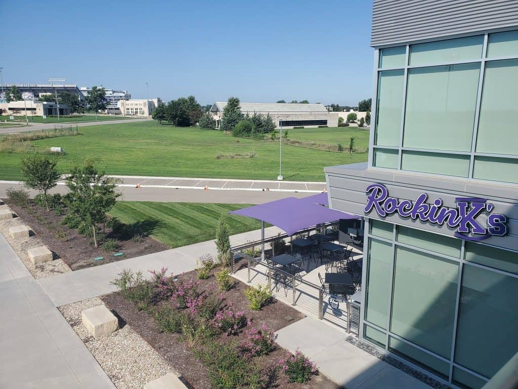 An exterior view of a modern building with large windows, featuring a restaurant named "Rockin' X's" with a patio covered by a purple awning. The peaceful outdoor seating area overlooks an open grassy field with some distant buildings. The sky is clear and blue.