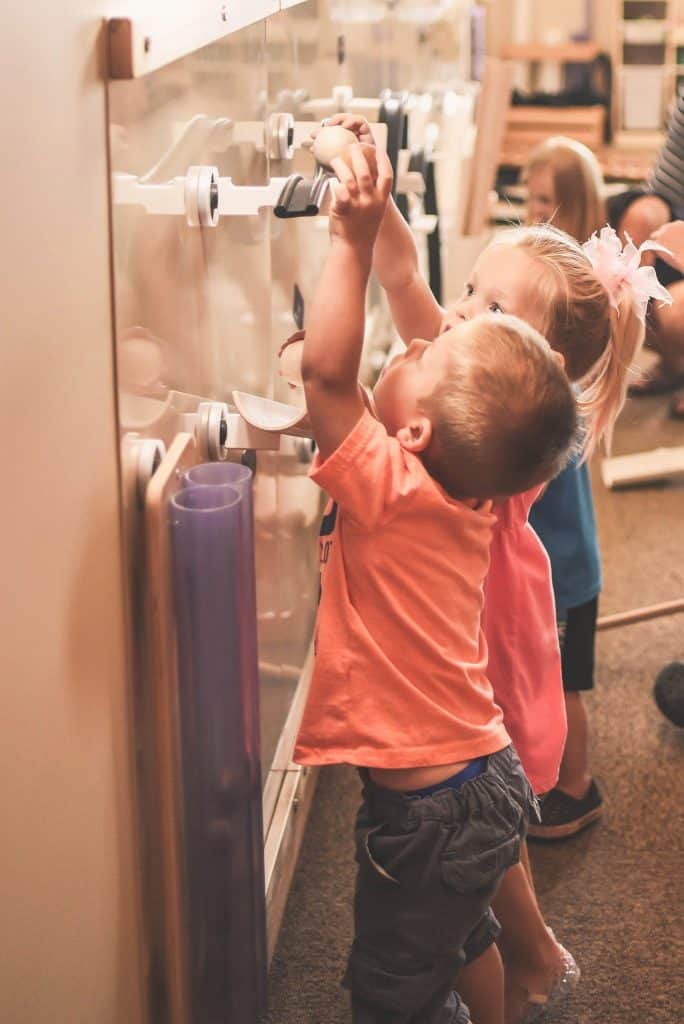 Two young children are playing with an interactive wall activity. The child in the foreground, wearing an orange shirt, is reaching up towards the wall. Another child, with a bow in their hair, watches and participates. The setting appears to be a classroom.
