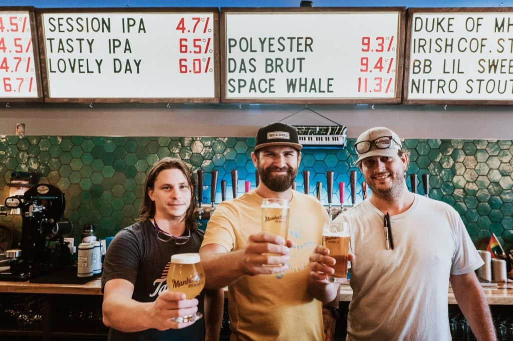 Three men standing in a bar, holding beers and smiling at the camera. Behind them, a beer menu on the wall lists different types of beers and their alcohol percentages. The bar has a green tiled wall and beer taps in the background.