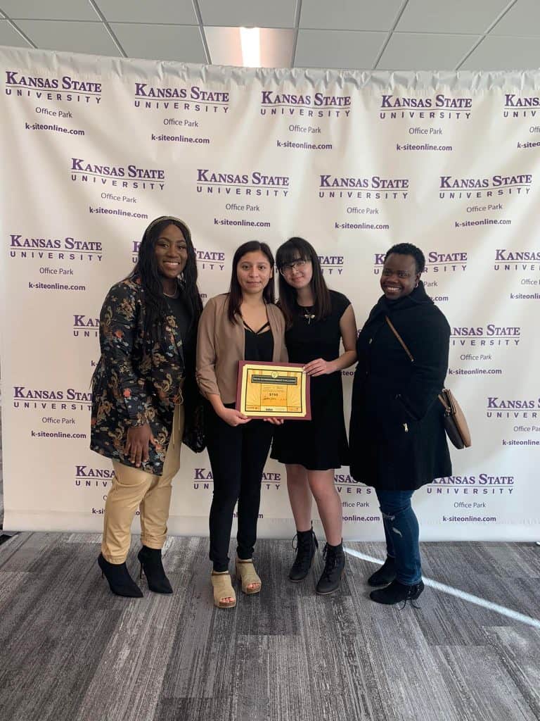 Four women are standing in front of a Kansas State University backdrop. The woman second from the left is holding a framed award. They are all smiling and dressed in smart casual attire.