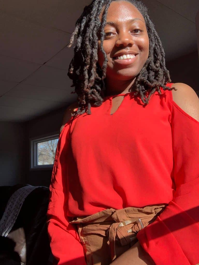 A person with shoulder-length twisted hair is smiling in a well-lit room. They are wearing a vibrant red blouse with cut-out shoulder details and brown trousers. Sunlight is streaming in from a window behind them, illuminating their face.