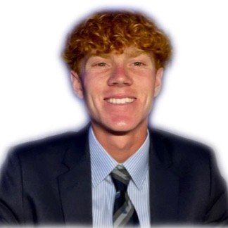 A young man with curly red hair is smiling. He is wearing a dark suit jacket, a light blue dress shirt, and a striped tie. The background is plain white, making his features stand out.