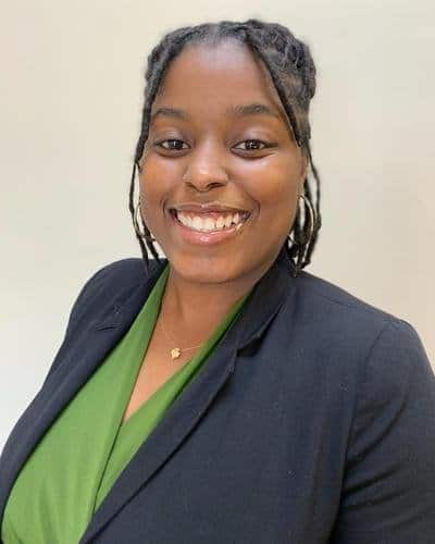 A woman with long braids smiles at the camera. She is wearing a black blazer over a green top and has a gold necklace. The background is plain and light-colored.