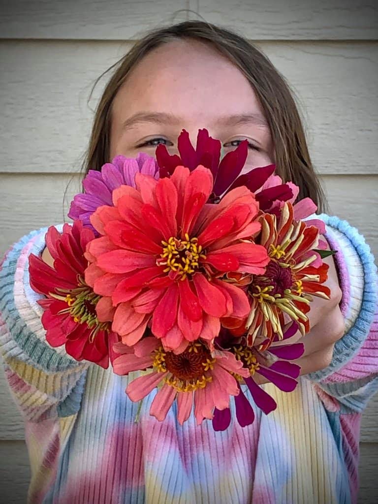 A person with long hair holds a vibrant bouquet of colorful zinnias, obscuring most of their face. They are wearing a tie-dye hoodie with shades of pink, blue, and white. The background features a beige wooden wall.