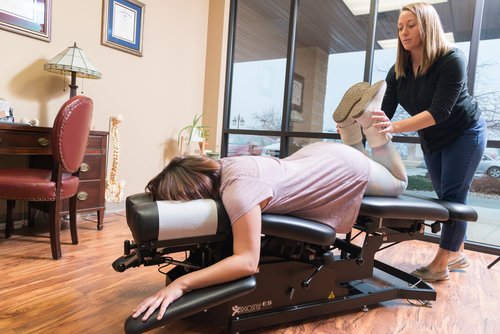 A woman lays face down on a chiropractic table while a female chiropractor, standing beside her, performs an adjustment on her leg. The room has wood flooring, a desk with a lamp, and a model of a spine resting on a cabinet.
