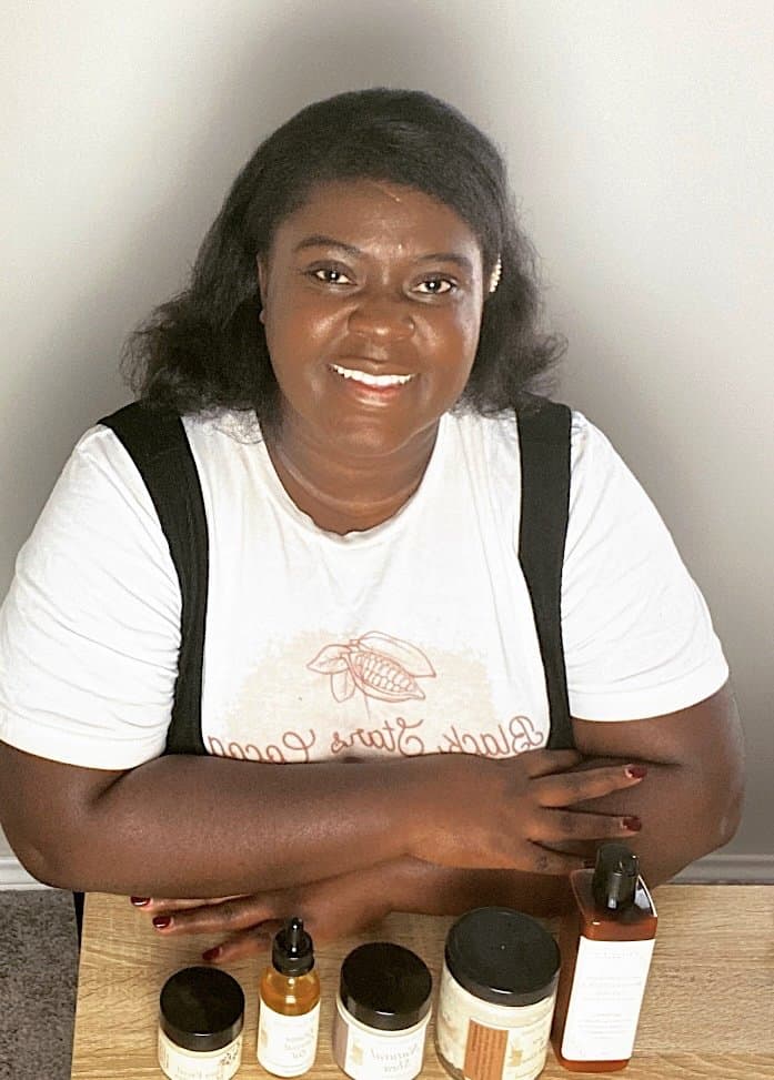 A person with long hair smiles warmly while seated at a table with various skincare products, including dropper bottles, jars, and a lotion bottle. They wear a white t-shirt and have their arms resting on the table.