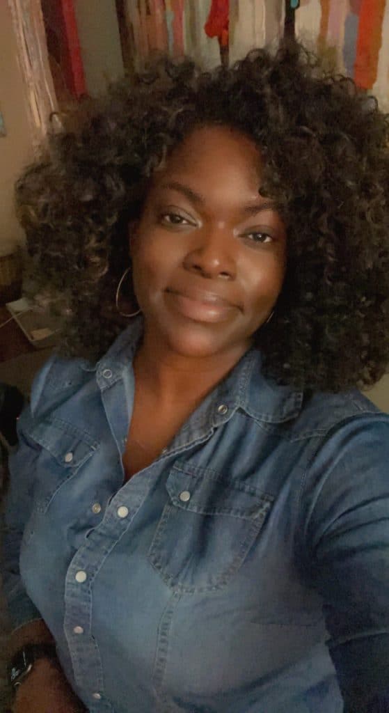 A person with curly hair wearing a denim shirt smiles at the camera while standing indoors. The background appears to have some colorful abstract art on the walls.