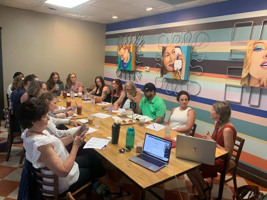 A group of people seated around a long table in a room with colorful striped walls and large portraits of women. They appear to be in a meeting or workshop, with some individuals looking at papers and others at laptops or phones.