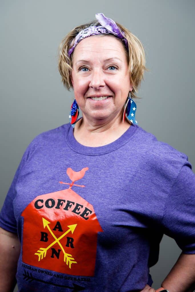 A person smiles at the camera, wearing a purple t-shirt with a bird and "Coffee BARN" printed on it. They also wear a purple headband and star-patterned earrings. The background is plain gray.