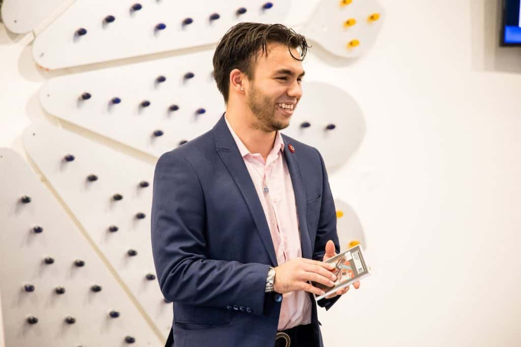 A man with dark hair, wearing a navy blazer and light pink shirt, stands smiling and holding a smartphone. Behind him is a wall decorated with white panels and small colorful hooks or pegs. The setting appears to be indoors, possibly a modern office or gallery.