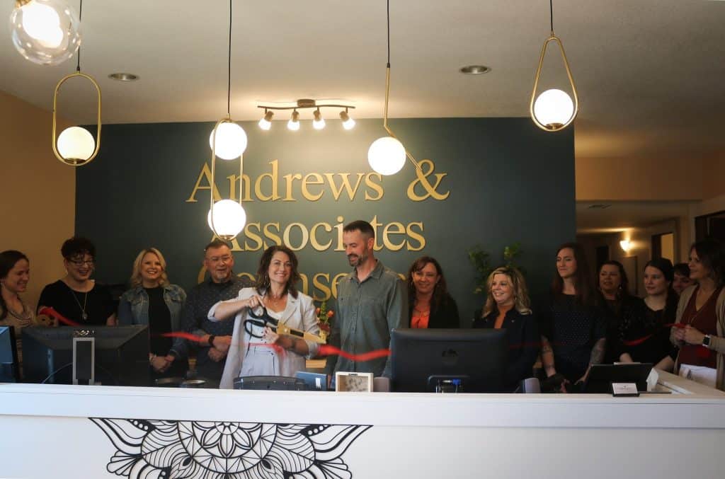 A group of people stand behind a reception desk with a sign that reads "Andrews & Associates." One woman cuts a red ribbon, holding large scissors. The room has pendant lighting and a decorative wall. Everyone is smiling and appears celebratory.
