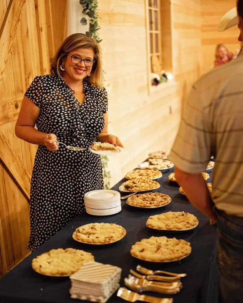 A smiling woman in a black and white polka dot dress serves herself a slice of pie at a table filled with an assortment of pies. The background features wooden walls and a partially visible person in a striped shirt standing in front of the table.