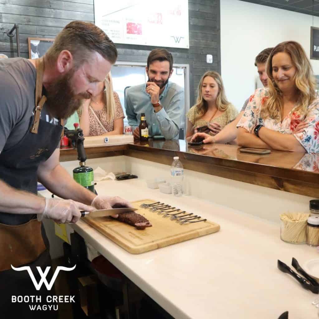 A chef slices Wagyu beef on a wooden cutting board while several people watch attentively from behind a counter. The setting appears to be a butcher shop or culinary demo area. A logo for Booth Creek Wagyu is visible in the bottom left corner of the image.