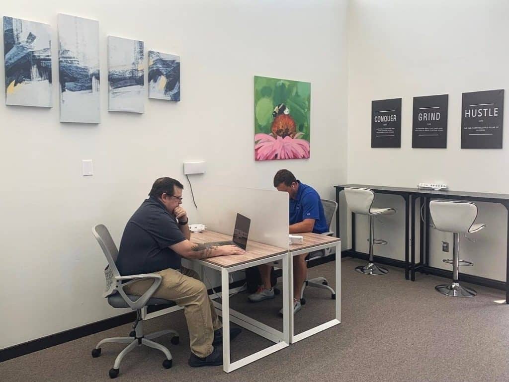 Two men sitting at separate desks working on laptops in a modern office space. The room features abstract wall art, motivational posters, and high-top seating along the back wall. A clear divider separates the men, and the atmosphere is professional and focused.