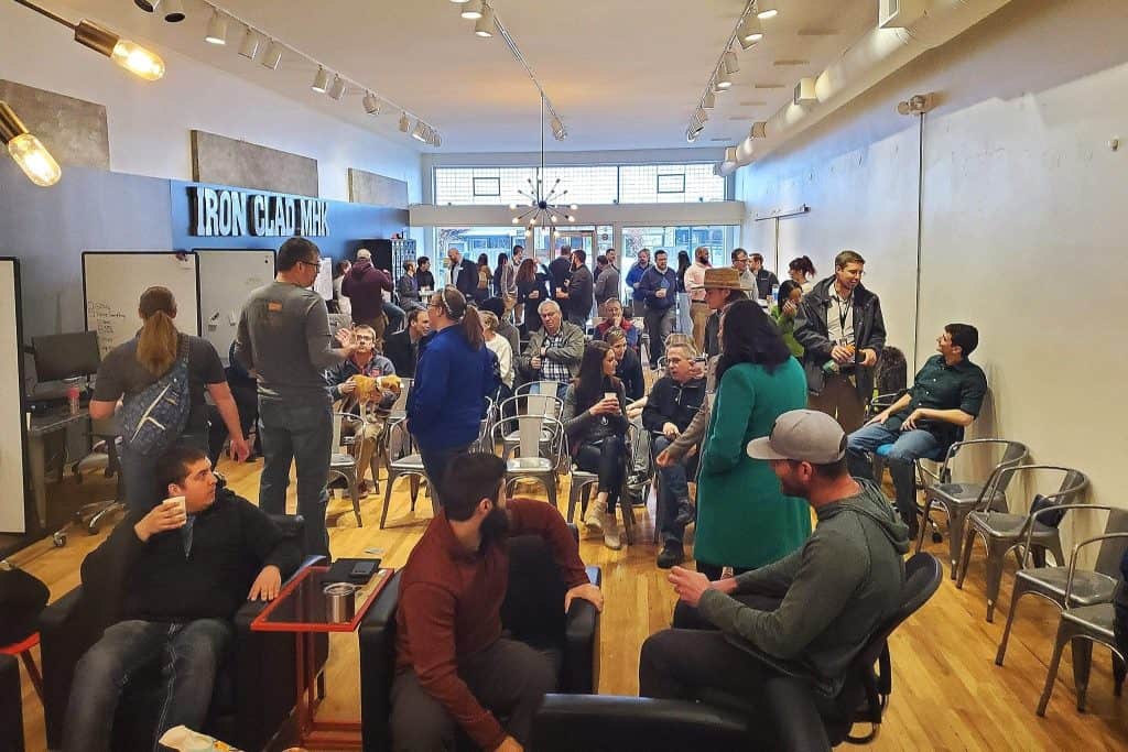 A crowded room with people seated and standing, talking and mingling. The space has wooden floors, high ceilings, and is brightly lit with modern fixtures. The wall has a sign that reads "IRON CAD MEN". One person is addressing the group in the background.