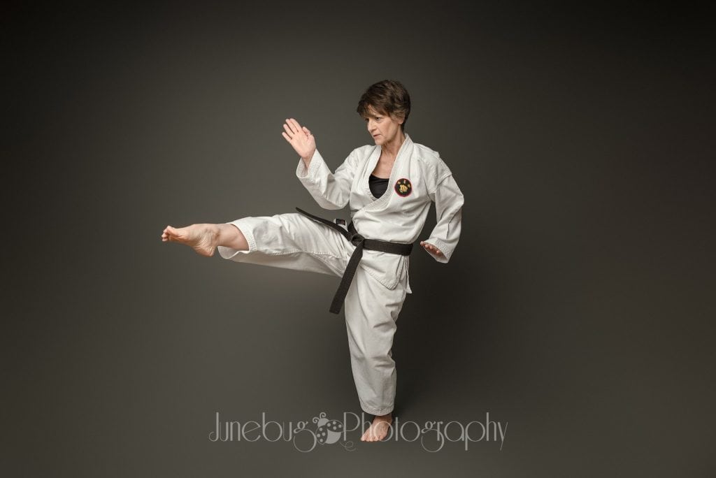 A person dressed in a white karate uniform with a black belt is performing a high kick. The individual is focused and poised, lifting one leg to the side while raising one arm in a defensive posture. The background is dark, and the image has a professional watermark.