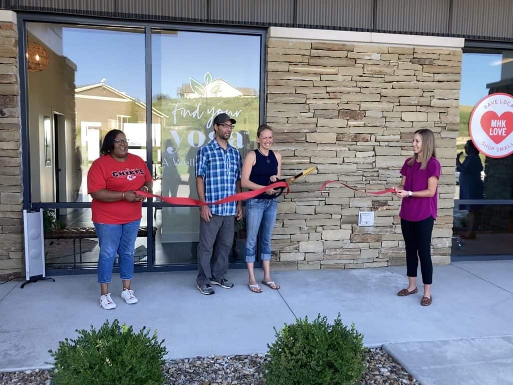 Four people stand in front of a building with "Find Your Joy" in the window, holding a large red ribbon and a pair of oversized scissors, preparing for a ribbon-cutting ceremony. The background features a stone wall and another window with "MHK Love" signage.