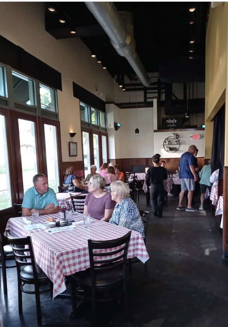 A casual restaurant with several people dining. The tables are covered with red and white checkered tablecloths. Large windows line one side, and a few more patrons can be seen standing near the entrance. The atmosphere is cozy and well-lit.