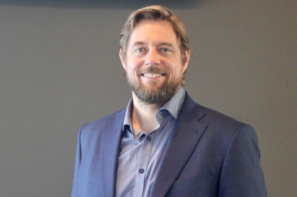 A man with light brown hair and a beard is smiling and wearing a blue blazer over a patterned shirt. He stands against a plain dark background.