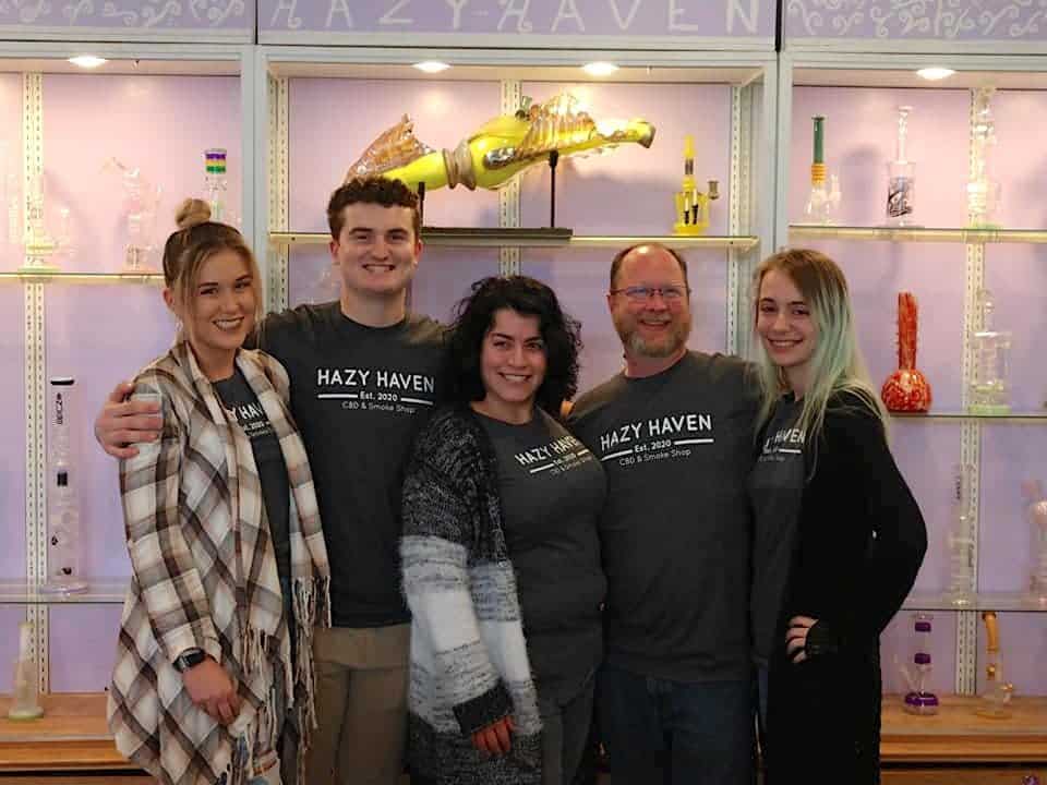 Five people are standing in a row, smiling at the camera. They are wearing "Hazy Haven" shirts and are in front of shelves displaying various glass items, including pipes and decorative pieces. The background has a decorative purple wall with "Hazy Haven" written at the top.