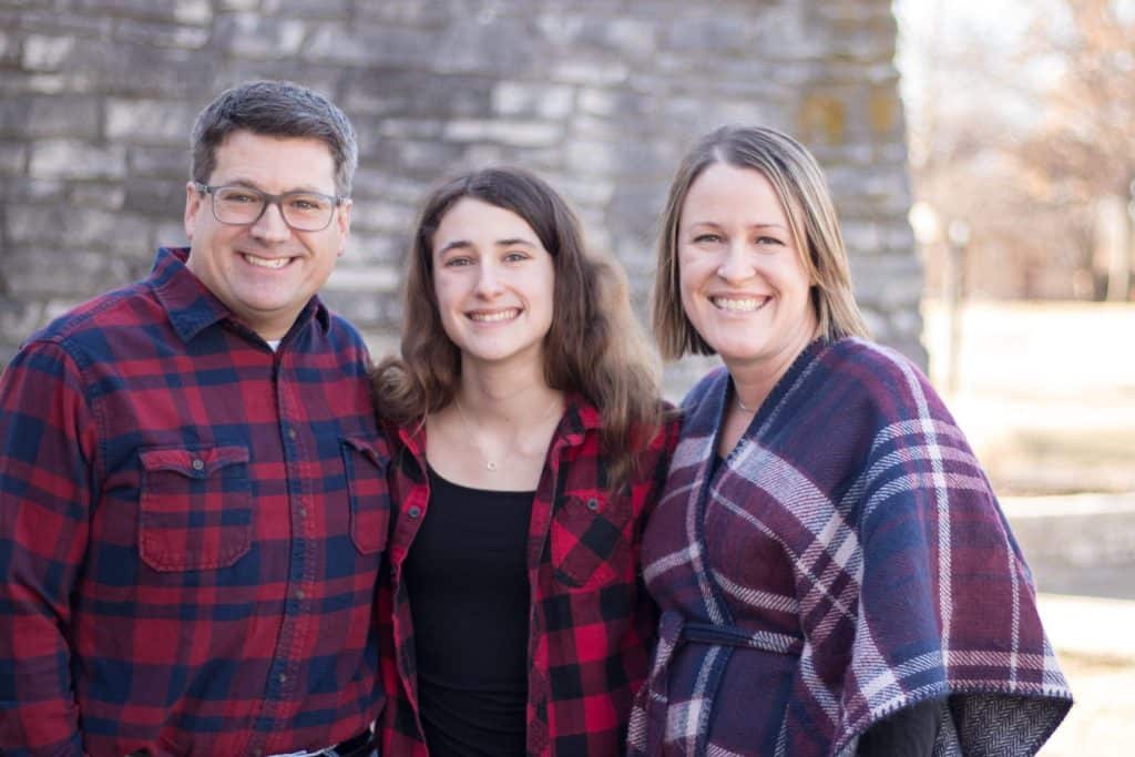 A smiling family of three stands outdoors. The father, wearing glasses and a red plaid shirt, stands on the left. The daughter, also in a red plaid shirt, is in the middle. The mother, in a red, blue, and white plaid shawl, stands on the right.