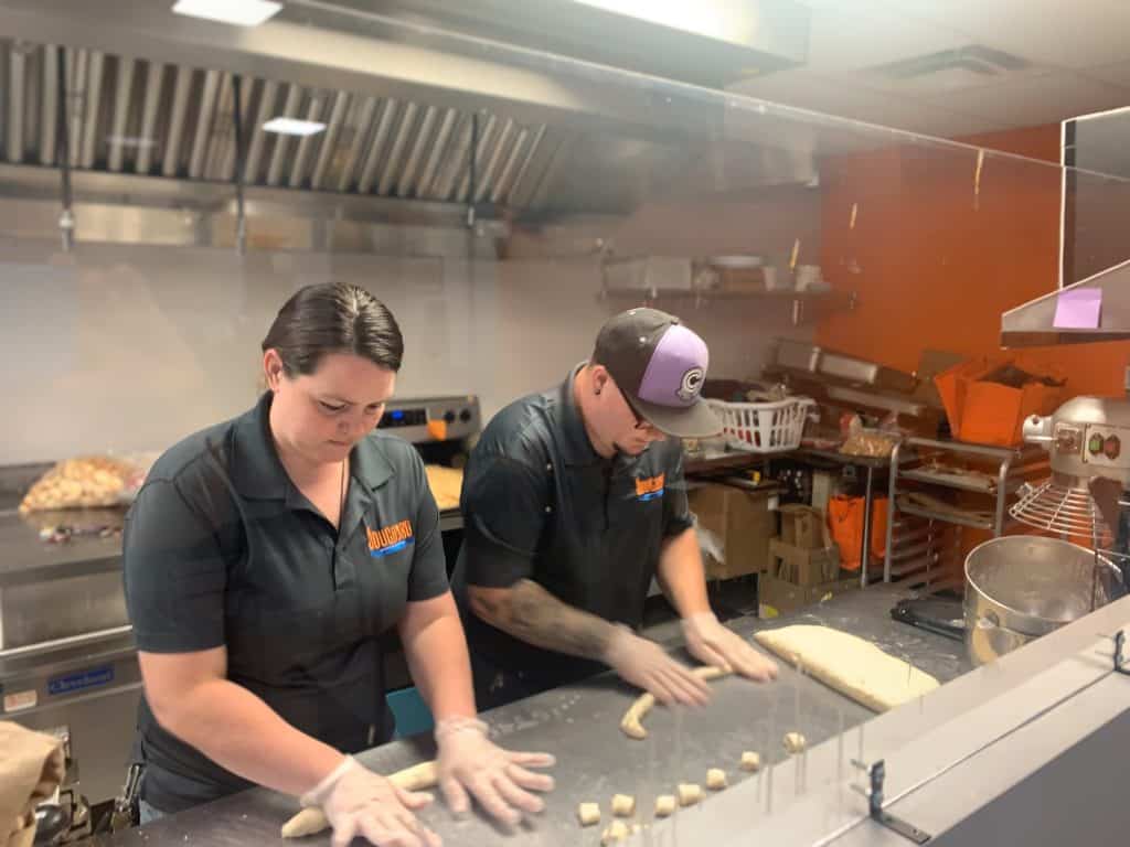 Two people are working together in a commercial kitchen, rolling out dough on a stainless steel counter. Both are wearing black uniforms and gloves. The kitchen is equipped with various utensils and baking supplies, and there is a protective screen in front of them.