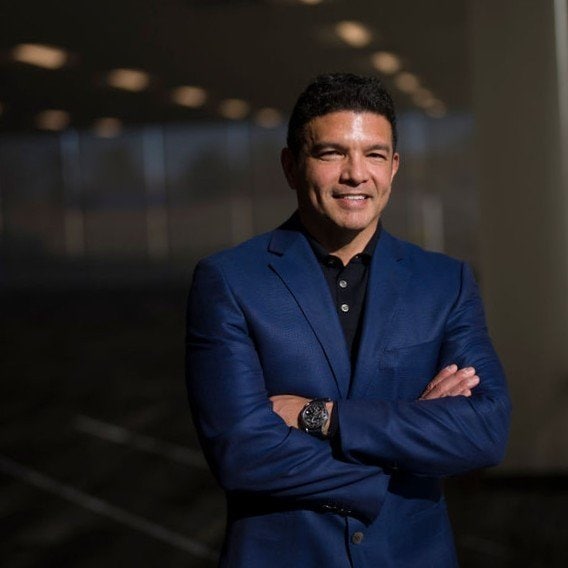 A man in a blue suit stands with his arms crossed and smiles. He wears a black shirt underneath the suit and a wristwatch on his left wrist, set against a dark, blurred background with soft lighting.