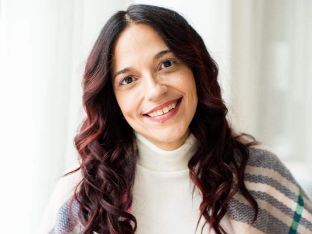 A woman with long, wavy, dark hair with red highlights, smiling at the camera. She is wearing a white turtleneck and a striped shawl. The background is softly blurred, suggesting an indoor setting with natural light.
