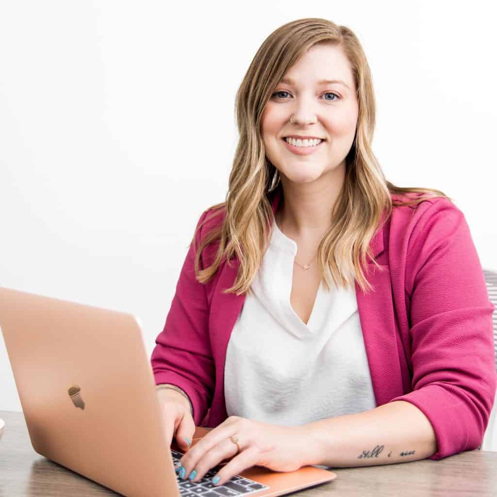 A smiling woman with long blonde hair is sitting at a desk working on a laptop. She is wearing a bright pink blazer and a white top. A tattoo is visible on her right forearm. The background is plain white.