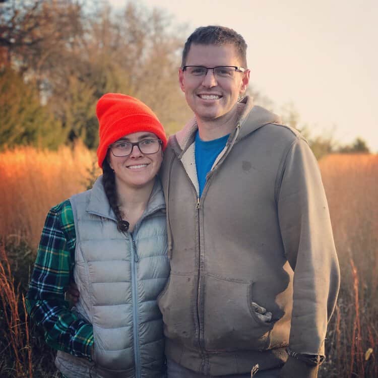 Two people stand close together in a sunlit field. The person on the left wears a bright orange beanie, glasses, a plaid shirt, and a puffy vest. The person on the right, who is taller, wears glasses, a blue shirt, and a hooded jacket. Both are smiling at the camera.