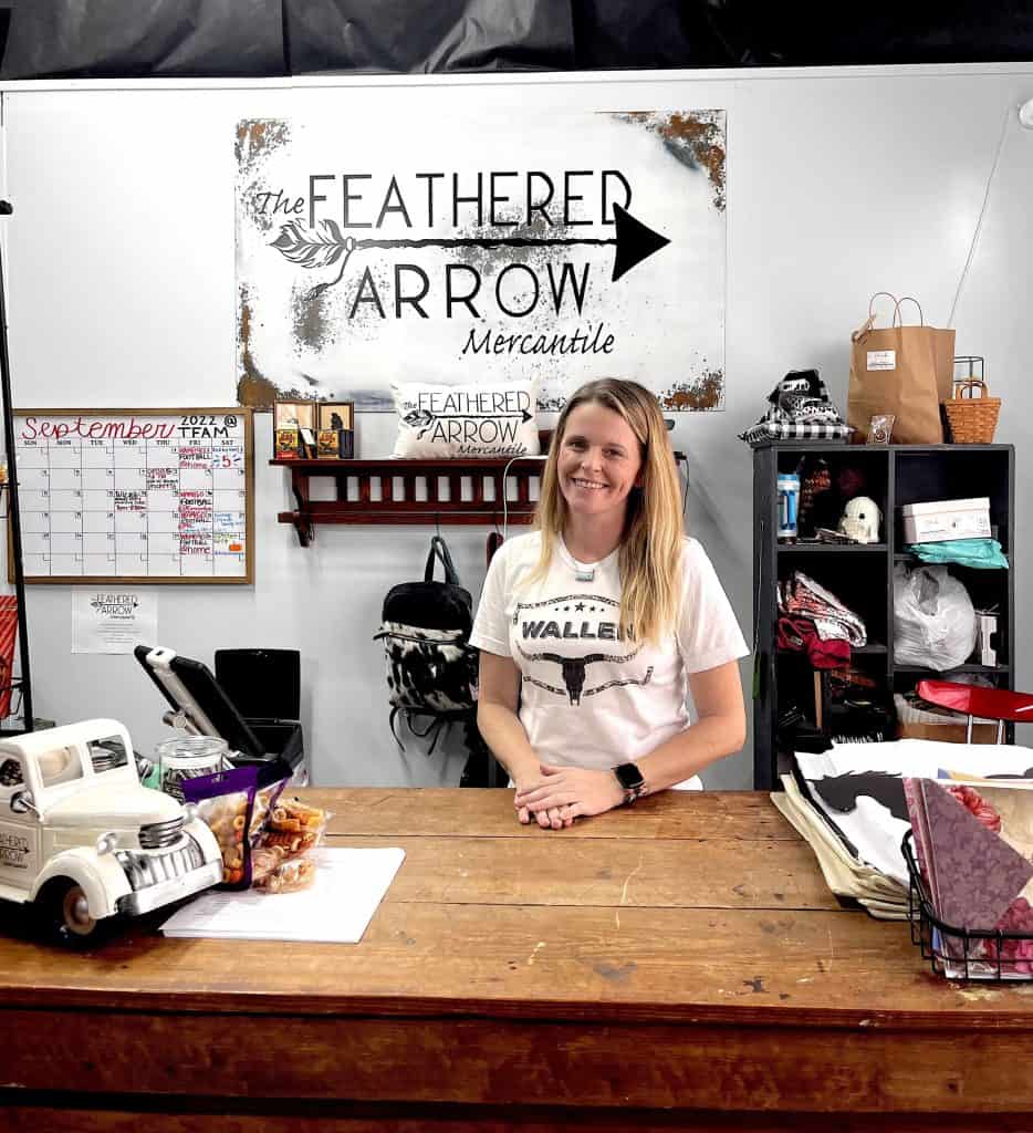A woman with long blonde hair stands behind a wooden counter at The Feathered Arrow Mercantile. The counter displays toy trucks, paper, and various other items. Shelves in the background hold bags, decor, and a calendar shows September. A sign with the store's name hangs above.