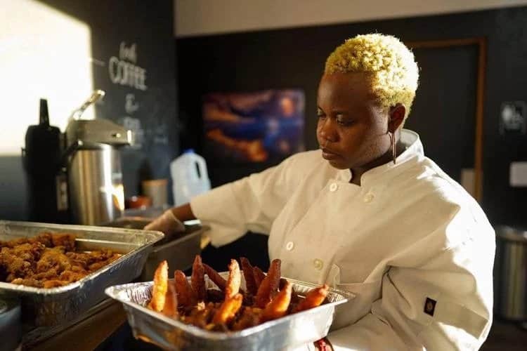 A person with short blonde hair, wearing a white chef coat, is arranging food on trays. The trays contain fried chicken and other food items. The person is in a kitchen with a coffee machine and a wall with writing in the background.