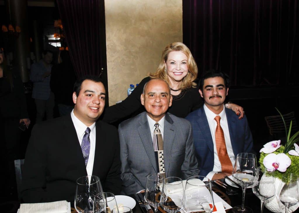 A group of four people is posed for a photo in a dimly lit restaurant. Three men in suits are seated at a table set with wine glasses and plates. A woman stands behind them, smiling. The table has an arrangement of flowers and napkins.
