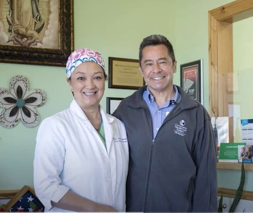 A smiling woman in a white lab coat and colorful scrub cap stands next to a smiling man in a gray zip-up jacket. They are indoors with a green wall, framed pictures, and certificates displayed in the background.