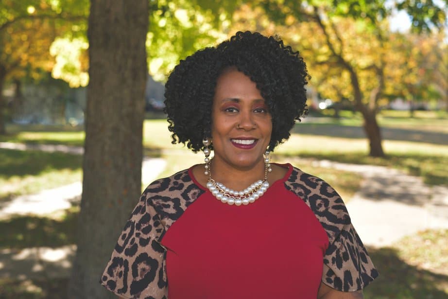 A woman with curly hair smiles at the camera while standing outdoors under a tree. She is wearing a red top with leopard-print sleeves and a pearl necklace. The background features sunlight filtering through green and yellow leaves.