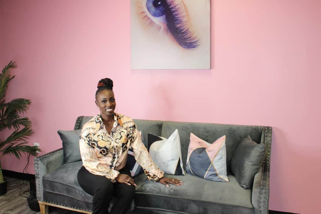 A woman sits on a gray couch with studded detailing, adorned with decorative pillows. She is wearing a patterned blouse and dark pants, and is smiling at the camera. Behind her, the wall is pink with a close-up image of an eye with long eyelashes. A plant is on the left.