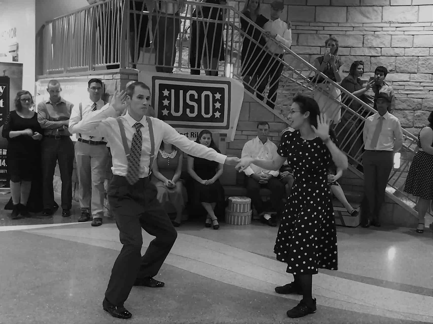 A man and woman swing dance in a public space, surrounded by onlookers sitting and standing near a staircase. The man wears a shirt and tie with suspenders while the woman wears a polka dot dress. A USO sign is visible in the background.