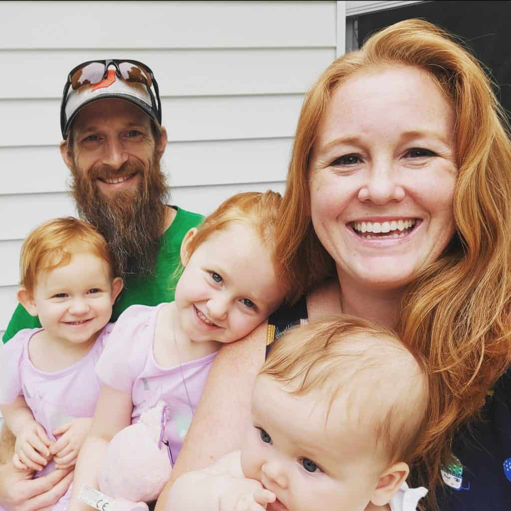 A family of five poses for a selfie. The father, with a beard and sunglasses on his head, is at the back. The mother, with long red hair, is in the front right. They are accompanied by three young children, two of whom have red hair and one with lighter hair. Everyone is smiling.