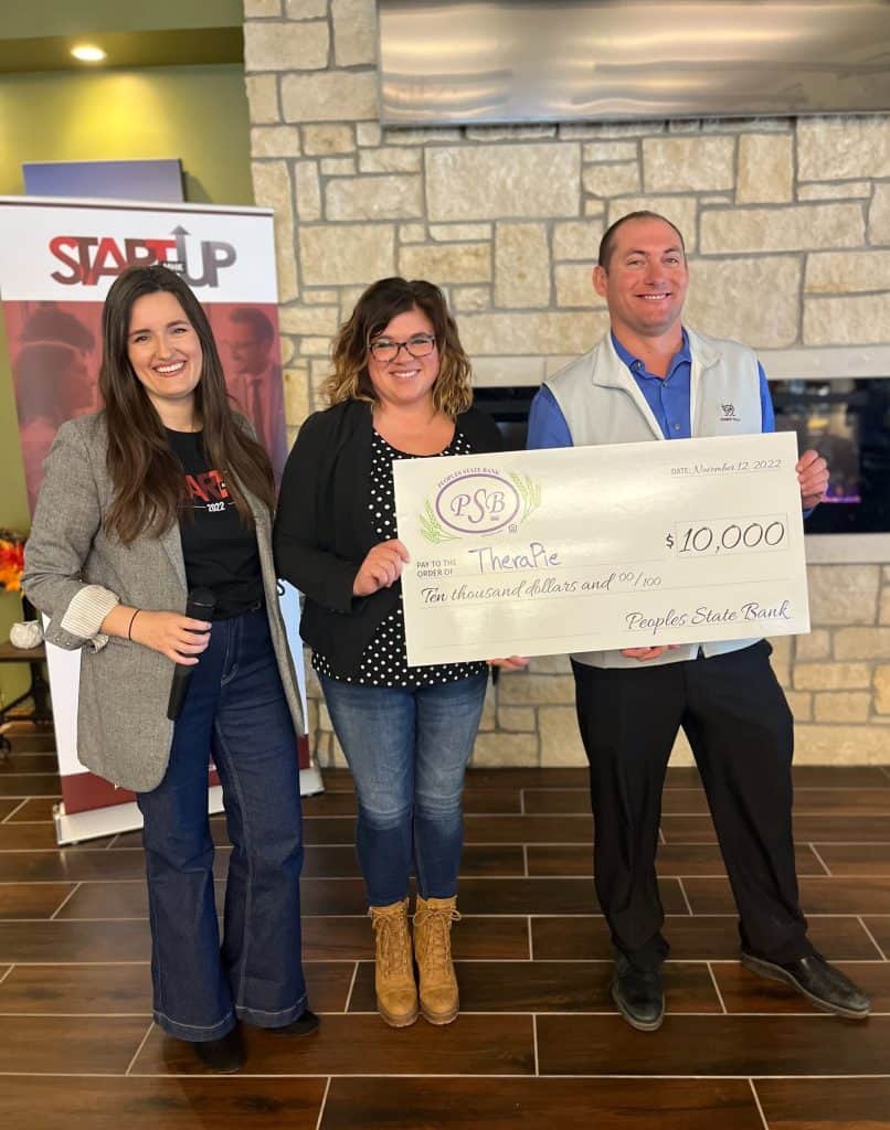 Three people stand indoors holding a large ceremonial check for $10,000 from Peoples State Bank to Therapie. They are smiling at the camera. Two of them are women, one in the middle holding the check, and a man is on the right also holding the check.