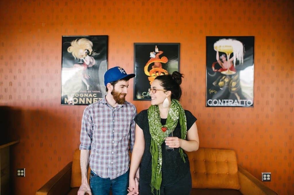 A man and woman are holding hands and smiling at each other. The man is wearing a blue cap and plaid shirt, and the woman has glasses, an updo, and a green scarf. They stand in front of a retro orange wall with three vintage posters. An orange couch is behind them.