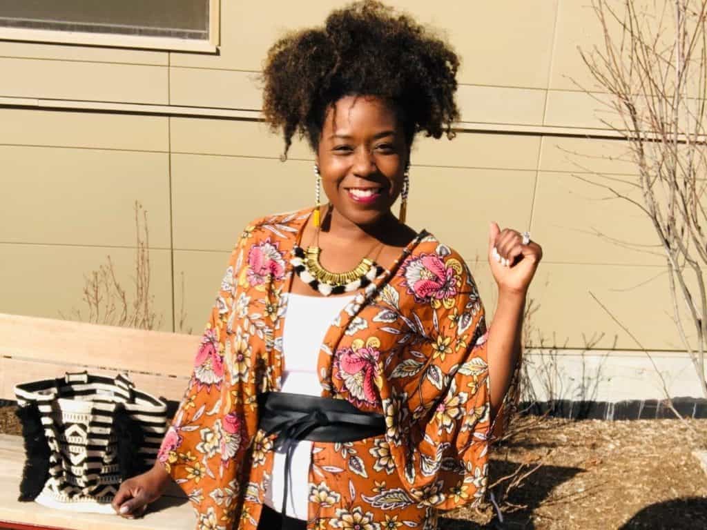 A woman with an afro hairstyle smiles and poses outdoors, wearing a floral kimono over a white top, accessorized with a chunky necklace and large earrings. She stands near a bench with a black and white patterned handbag beside her. The background has a beige building.