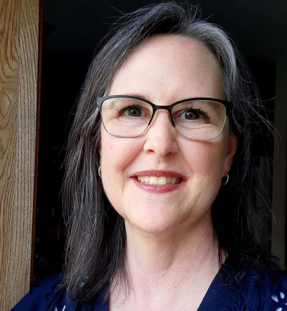 A woman with shoulder-length gray hair and glasses smiles at the camera. She is standing indoors near a wooden doorframe, wearing a navy blue top. The background is softly lit and out of focus.