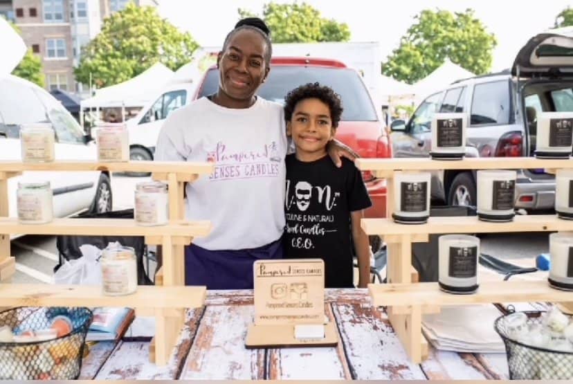Two people stand behind a candle display at an outdoor market. The adult on the left wears a white shirt with text, and the child on the right wears a black shirt with text. The wooden shelves hold various candles in jars. Cars and tents are visible in the background.