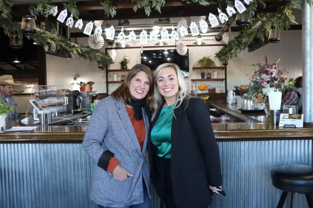 Two women are standing and smiling in front of a rustic bar adorned with hanging greenery and a small white house garland. One is wearing a blue striped blazer; the other is in a green blouse and black jacket. The background shows bar counters, patrons, and floral arrangements.