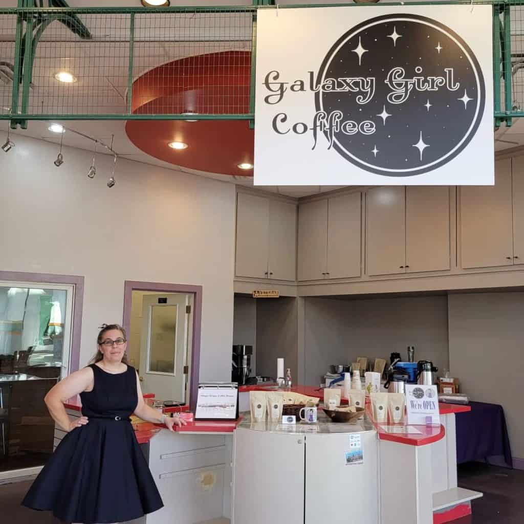 A woman in a black dress stands behind a counter in a coffee shop named Galaxy Girl Coffee. The counter displays various coffee bags and a sign that says "We're OPEN". A large circular sign with the coffee shop's name and a starry design hangs above.