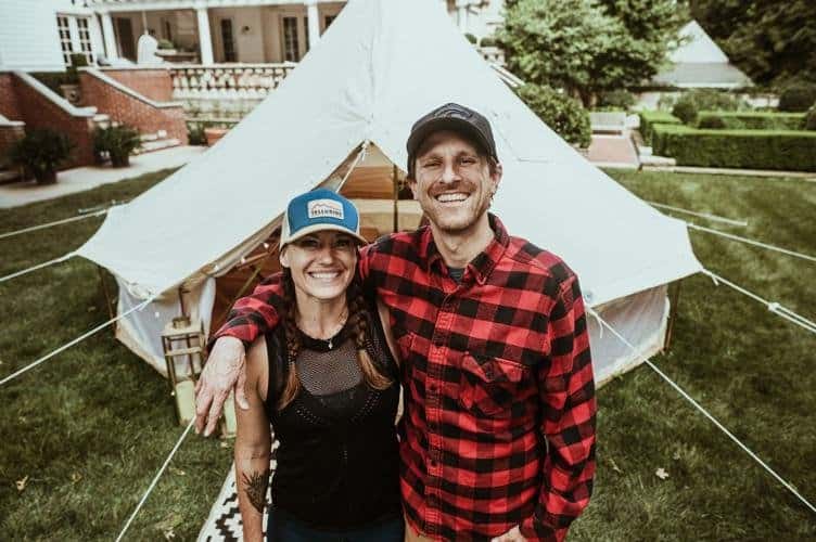 A smiling couple stands in front of a large, canvas tent set up on a lawn. The woman is wearing a blue baseball cap and a black mesh top, and the man is wearing a black cap and a red plaid shirt. The background features a well-manicured garden and a large house.