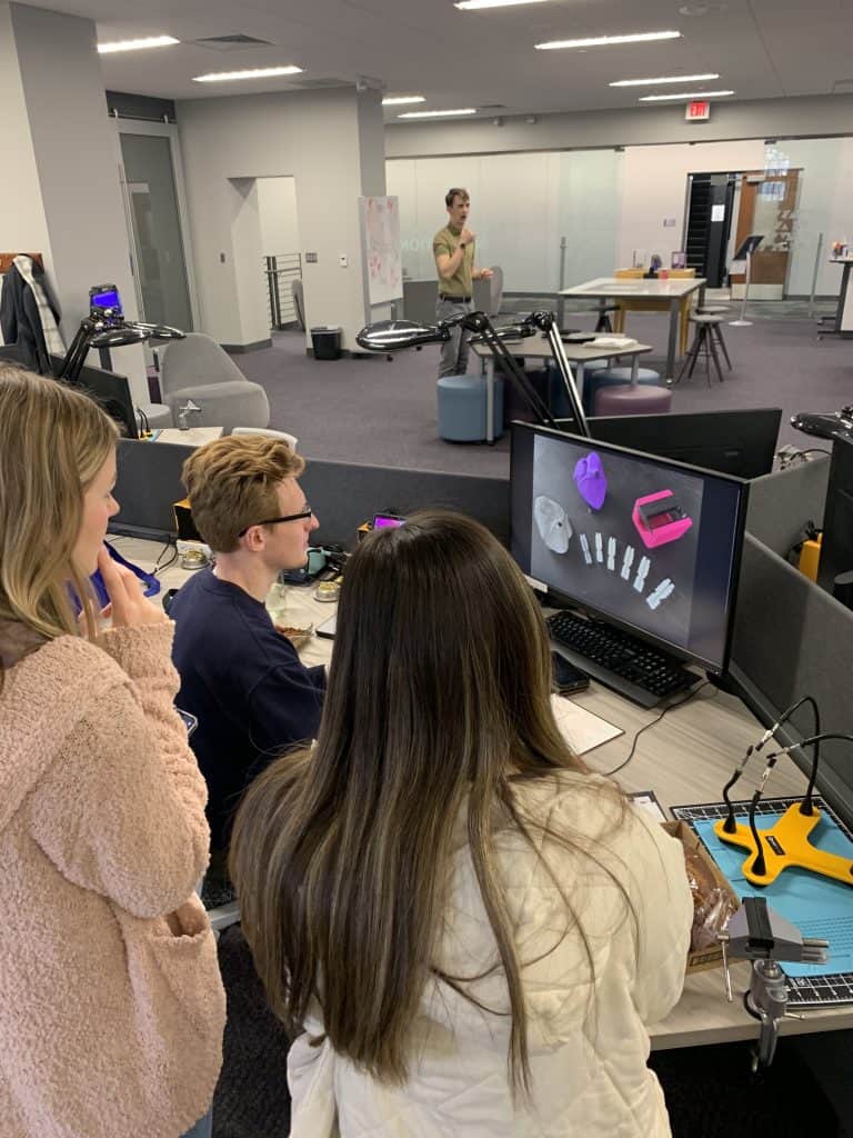 Three people are gathered around a computer desk, observing or discussing the 3D design displayed on a monitor. The office space has 3D printers and workstations. Another person stands in the background near a whiteboard and gestures while speaking.